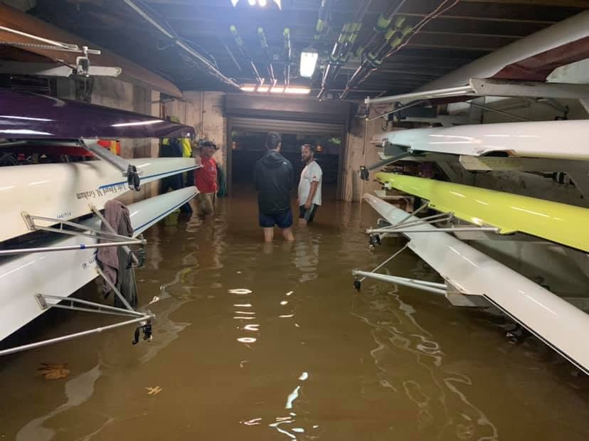 Muddy Schuylkill River flood water standing knee-deep in the Crescent Boat Club boat bay, with members wading between racked shells